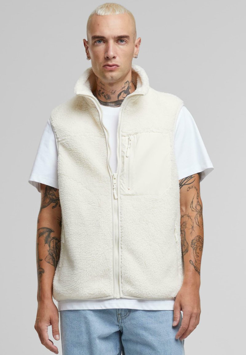 Gilet en polaire couleur crème avec un col montant, fermeture éclair et poche avant. Porté sur un t-shirt blanc, il présente une texture douce.