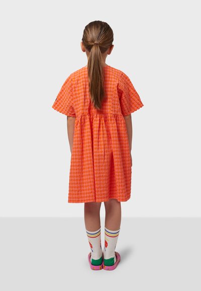 Orange, rutigt flickklänning med korta ärmar och samlad midja. I kombination med vita strumpor med mönster och rosa sandaler.