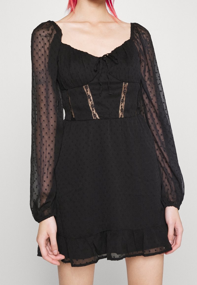 Black short dress with puffed sheer sleeves, dotted texture, a fitted bodice, lace accents, and a ruffled hemline. Features a tie front detail.