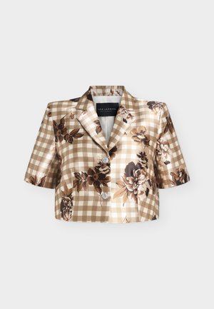 Femme portant un chemisier à manches courtes fleuri à carreaux, pantalon marron taille haute, talons crème, tenant un sac à main beige, debout devant un fond blanc.