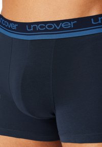 Close-up van een persoon die marineblauwe boxershorts draagt met een blauwe tailleband waarop "uncover" staat.