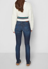 Jeans ajustados azules con bolsillos traseros, combinados con un suéter de punto trenzado color crema que presenta una cinturilla a rayas. El calzado tiene una suela marrón.