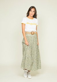 T-shirt blanc avec des bordures jaunes et le texte "HEUREUX", associé à une jupe longue florale verte et une ceinture beige. Des baskets blanches complètent la tenue.