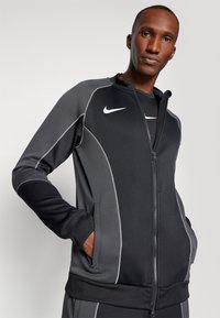 Nike užtrauktukas striukė juodos ir pilkos spalvos. Ypatybės: kontrastingas siuvinėjimas, stovinti apykaklė ir šoninės kišenės. Šiek tiek aptemptas siluetas su sportiniu dizainu.