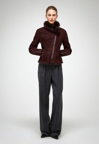 Mujer de pie con chaqueta de ante marrón oscuro con cuello de piel y pantalones anchos grises, sobre un fondo liso claro.