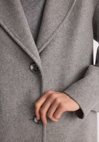 Zapinanie szarego wełnianego płaszcza z dużymi, dopasowanymi guzikami, z naciskiem na szczegóły ręki i przodu płaszcza.