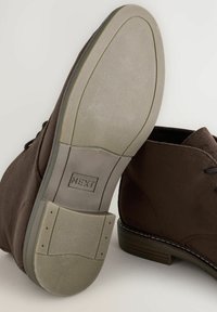 Botas ankle de ante marrón mostradas desde el lado y la parte inferior, destacando la suela texturizada con la marca "NEXT" y los cordones.