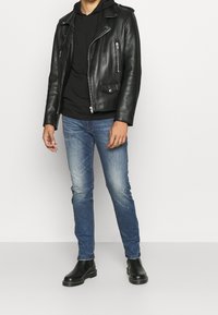 Chaqueta de cuero negra estilo motorista con cremalleras plateadas, llevada sobre una sudadera con capucha negra. Combinada con vaqueros ajustados azul claro y botines negros.