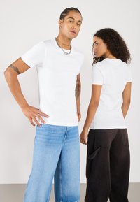 Hvide ribbede t-shirts båret af to personer, kombineret med lys blå og mørkebrun løse jeans. Enkel rund halsdesign, korte ærmer.