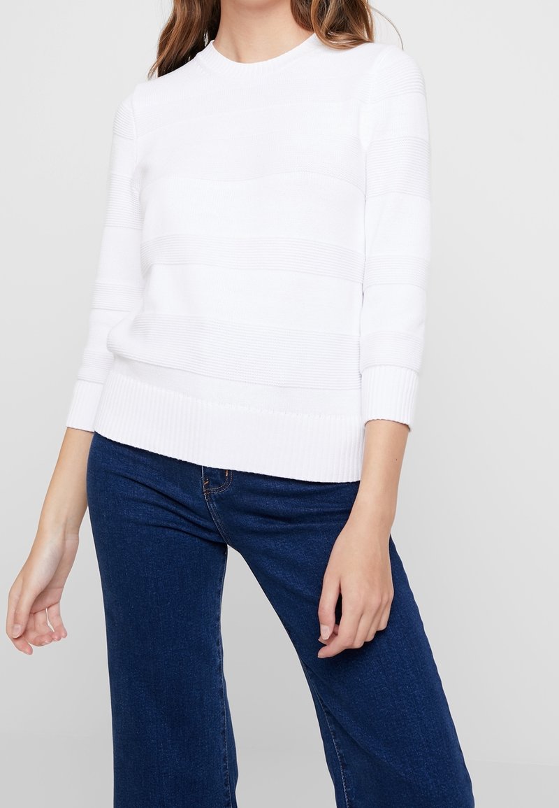 Weißer Rippstrick-Pullover mit horizontalem Streifenmuster, Rundhalsausschnitt und langen Ärmeln, kombiniert mit dunkelblauen weiten Jeans.