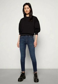 Jovem mulher de pé, vestindo um suéter preto oversized, jeans skinny azuis e botas pretas de plataforma com cadarço, contra um fundo cinza liso.