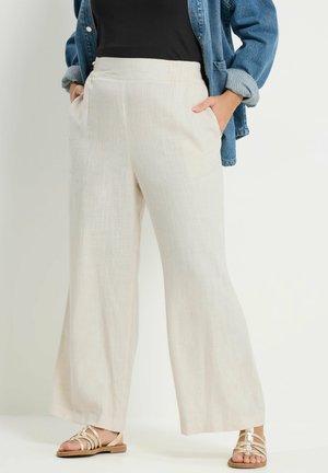 Kvinde iført cremelin bukser med vide ben og hænderne i lommerne, kombineret med en sort top og blå denimjakke, stående med sandaler.