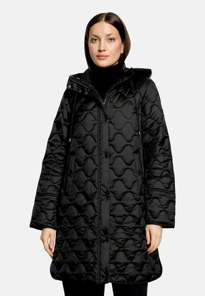 Frau trägt einen langen schwarzen gesteppte Steppmantel mit Kapuze und vorderer Knopfleiste über einem schwarzen Rollkragenpullover.