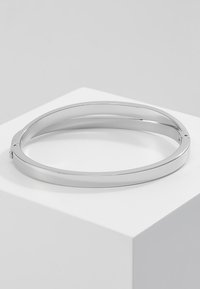 Brățară din metal argintie cu o suprafață netedă și lustruită și o formă ovală elegantă. Dispune de un balama subtilă pentru închidere.