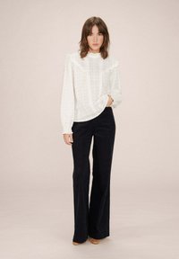 Blusa bianca con ruches, motivo floreale e maniche lunghe, abbinata a pantaloni neri a zampa d'elefante e tacchi beige. Sfondo neutro.