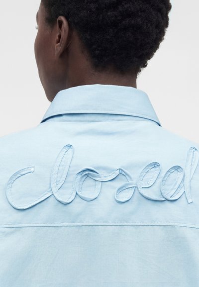 Άτομο με κοντά σγουρά μαλλιά γυρισμένο προς τα πίσω, φοράει ένα ανοιχτό μπλε πουκάμισο με κεντημένο κυματιστό κείμενο στην πλάτη.