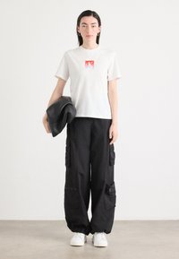 Witte katoenen t-shirt met een rood en oranje gradientlogo, gecombineerd met losse zwarte cargobroeken met meerdere zakken en ritsen. Witte sneakers.