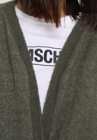 Cardigan lavorato a maglia verde oliva con design a V, indossato sopra una maglietta bianca con testo audace in nero. Tessuto morbido e vestibilità ampia.