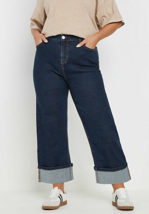 Jeans a gamba larga di colore blu scuro con vita alta, caratterizzati da cuciture a contrasto e risvolti di denim in una tonalità più chiara; abbinati a sneakers bianche.