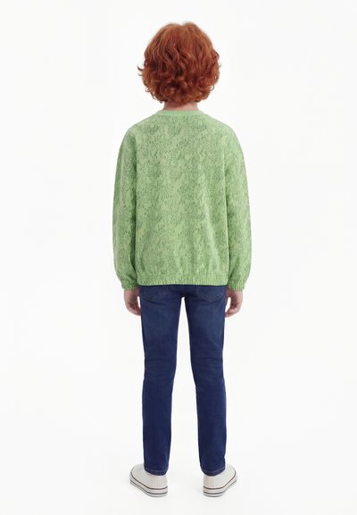 Barn med krøllete rødt hår står med ryggen mot, iført en lys grønn mønstret genser, mørkeblå jeans og hvite joggesko.