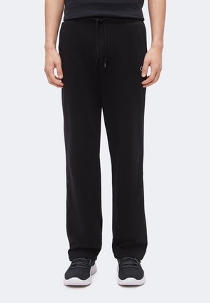 Personne portant un pantalon de survêtement noir à cordon, un haut noir et des baskets noires avec semelles blanches, debout devant un fond uni.
