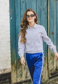 Gestreept overhemd met knopen in wit en blauw, gecombineerd met blauwe sportieve broek met witte zijstrepen. Lang, golvend haar en een zonnebril.