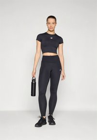 Mustast lõhikuga sportlik topp ja kõrge vöökohaga retuusid venivast kangast. Mustad spordijalanõud jalas ja musta veepudelit hoides.