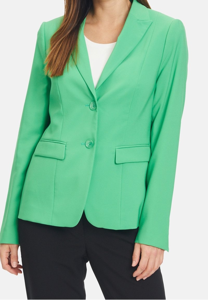 Veste verte en tissu lisse, dotée d'un col cranté, de deux boutons et de deux poches avant. Associée à un haut blanc en dessous.