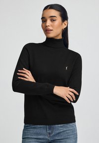 Kobieta o długich ciemnych włosach, ubrana w czarny sweter z golfem z małym wyhaftowanym logo polo zawodnika oraz niebieskie dżinsy, z założonymi skrzyżowanymi ramionami.