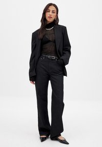 Black blazer over a sheer black turtleneck, paired with high-waisted black jeans and classic black flats. Features a silver necklace and a belt.