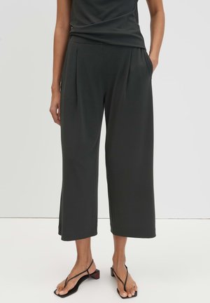 Donna con pantaloni neri a palazzo corti con tasche e sandali neri con tacco basso e cinturini, in piedi su pavimento bianco.