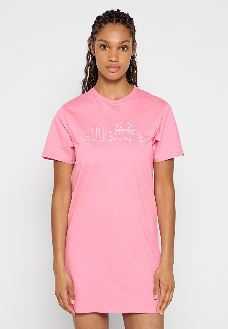 Mujer con el cabello trenzado, vistiendo un vestido rosa de manga corta con el logo blanco de "ellesse", de pie frente a un fondo blanco liso.