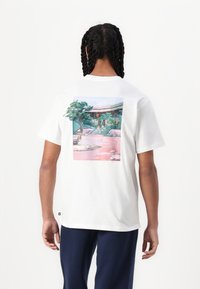 Weißes Baumwoll-T-Shirt mit einer bunten Illustration auf der Rückseite, die eine Landschaft mit Bäumen und einem Gebäude zeigt, sowie einem kleinen Logo.