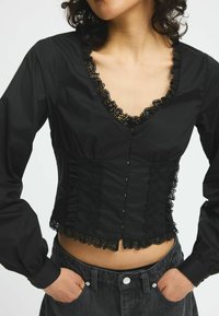 Blusa nera con scollo a V e rifiniture in pizzo. Presenta maniche lunghe voluminose e un busto aderente con bottoni decorativi e bordi ondulati.
