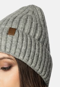 Beanie grigia a maglia con texture a coste. Presenta un piccolo tag in pelle marrone sul lato. Design a cupola affusolata. Materiale morbido e caldo.