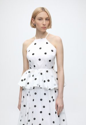 Mujer con cabello rubio corto que lleva un vestido halter blanco con lunares negros y una cintura peplum sobre un fondo blanco liso.