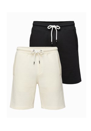 Homme portant un t-shirt graphique blanc et un short noir, debout avec une main dans la poche sur un fond uni.