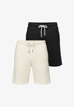 Mann trägt weißes Grafik-T-Shirt und schwarze Shorts, steht mit einer Hand in der Tasche vor einfarbigem Hintergrund.