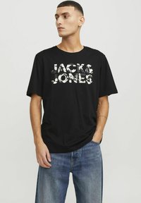 Juoda medvilninė marškinėliai su balta tekstūrine "JACK & JONES" logo, derinami su šviesiai mėlynomis džinsais, sukuriantys kasdienišką įvaizdį.