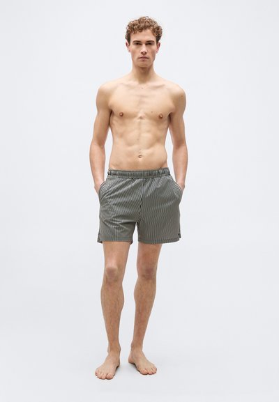 Jeune homme debout pieds nus, les mains dans les poches, portant un short à rayures verticales noires et blanches sur un fond clair uni.