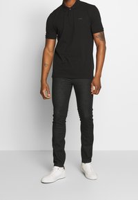 Camiseta polo negra de algodón con corte slim, mangas cortas y un cierre de tres botones, combinada con jeans oscuros y zapatillas blancas.