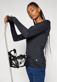 Black long-sleeve top made of stretchy material, featuring a round neckline and logo at the hem. The model holds a snowboard with bindings.