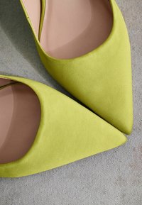Chaussures en suède vert à bout pointu avec une texture lisse et un décolleté, révélant une semelle insérée nude sur une surface grise.