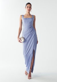 Kobieta w bezrękawowej lawendowej wieczorowej sukni z bocznym rozcięciem, trzymająca białą torebkę i nosząca białe szpilki oraz perłowe kolczyki.
