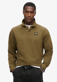 Olivgrünes Langarm-Polohemd mit Kragen, drei Knöpfen und einem Logopatch. Verfügt über einen strukturierten Stoff und gerippte Bündchen.