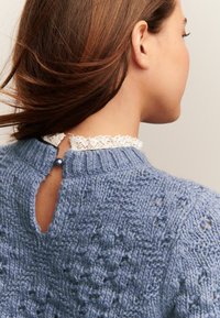 Mujer con cabello castaño lleva un suéter de punto texturizado azul con cierre de botón de perla y un collar de encaje blanco en la parte posterior.
