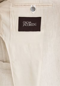 Béžová látka s hladkým povrchem, s hnědou etikou s logem "OSCAR JACOBSON", s uzávěrem na knoflík a ševovým detailem.