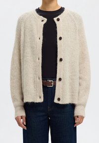 Person som bär en beige fluffig stickad cardigan med mörka knappar över en svart skjorta, mörka jeans och ett brunt bälte, med avslappnade händer vid sidorna.