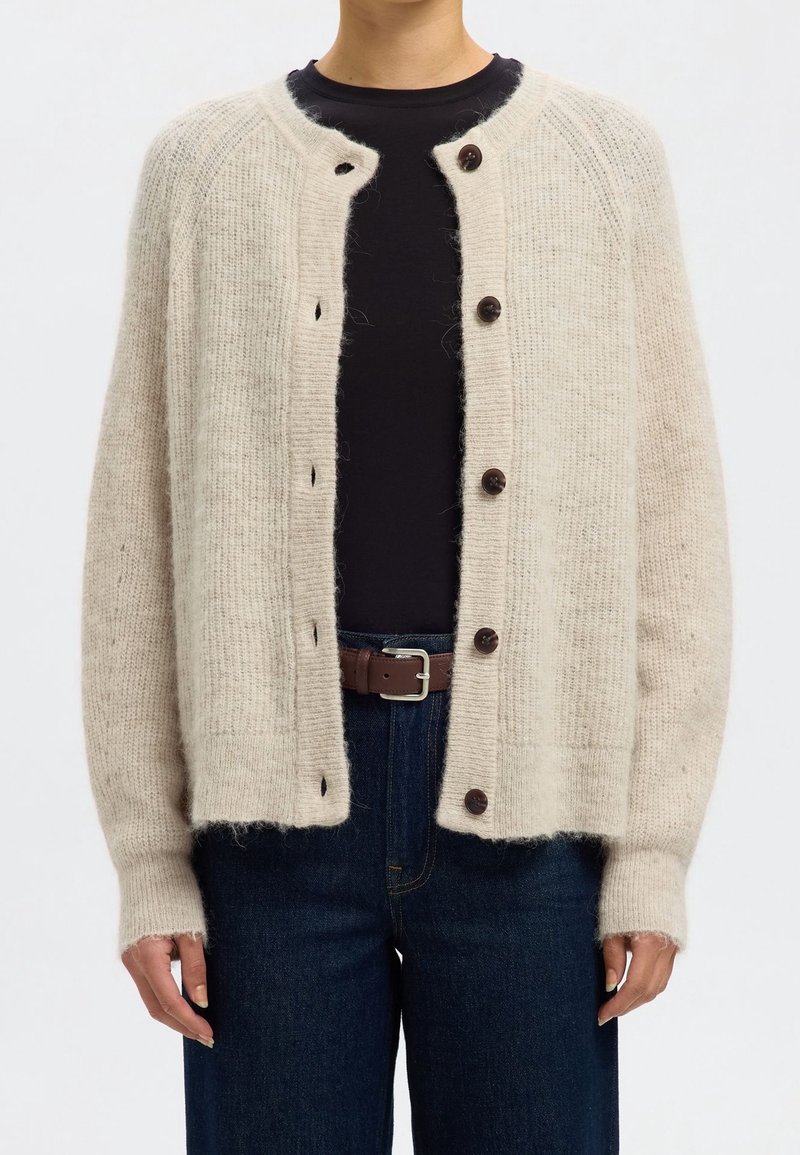 Person som bär en beige fluffig stickad cardigan med mörka knappar över en svart skjorta, mörka jeans och ett brunt bälte, med avslappnade händer vid sidorna.