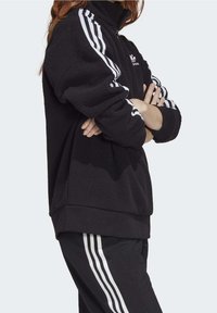 Svart fleece zip-up jacka med horisontella svarta och vita ränder på ärmarna. Har en hög krage och avslappnad passform.